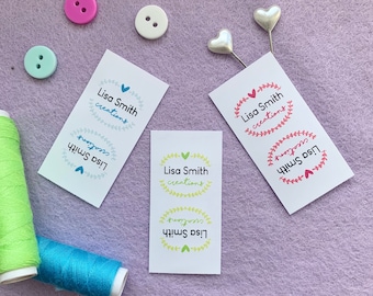 Op de afbeelding: Drie witte visitekaartjes met de tekst "Lisa Smith creations" in een kransontwerp. De kransen zijn in verschillende kleuren: blauw, groen en roze. De kaartjes liggen op een paarse ondergrond met knopen en spoelen garen.