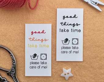 May include: Two white care labels with the text "good things take time" and a hand washing symbol. The text is in a handwritten font and the labels are on a beige background.