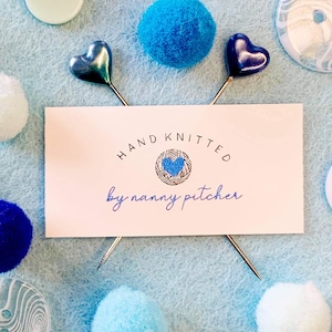 May include: A white card with the text "HAND KNITTED by nanny pitcher" in black and blue lettering. The card is surrounded by blue and white pom-poms and two silver heart-shaped pins.
