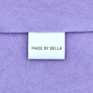 以下が含まれることがあります： 紫色の布に「MADE BY BELLA」と黒文字で書かれた白いラベルが付いている。
