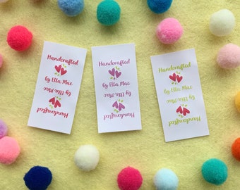 Op de afbeelding: Drie witte rechthoekige labels met de tekst "Handcrafted by Ella Mac" en een roze hart met groene bladeren. De labels liggen op een gele ondergrond met kleurrijke pompoenen.