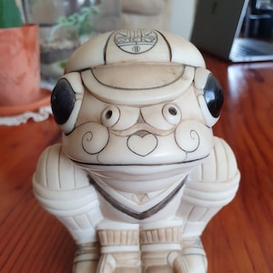 Könnte beinhalten: Eine weiße Keramikfigur eines Frosches, der ein Cricket-Uniform trägt. Der Frosch trägt eine weiße Kappe mit dem Wort "WICK" darauf und hat einen schwarzen Schnurrbart und Augenbrauen. Der Frosch hält einen Cricket-Schläger in seiner rechten Hand und einen Cricket-Ball in seiner linken Hand.