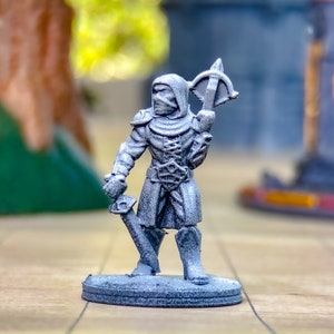 May include: A miniature figurine of a hooded warrior, holding a crossbow and a sword. The figure is gray and stands on a circular base. The background is blurred, suggesting a tabletop gaming environment.