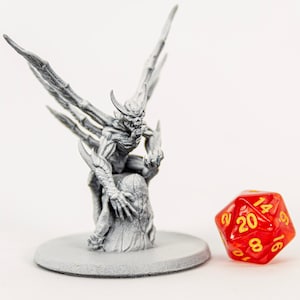 May include: A gray, detailed miniature of a demon with wings, sitting on a rock, next to a red 20-sided die with yellow numbers.