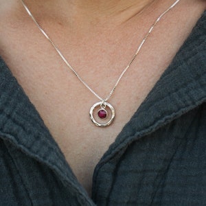 May include: A silver chain necklace with a small, hammered circle pendant featuring a red gemstone.