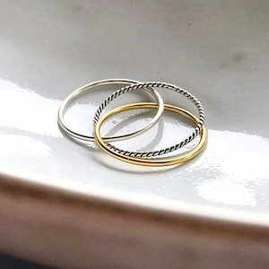 May include: Three thin rings stacked on top of each other. The top ring is gold, the middle ring is silver, and the bottom ring is silver with a twisted design.