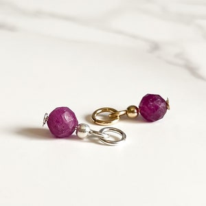 May include: Two purple gemstone beads with gold and silver jump rings. The beads are faceted and have a shiny finish.