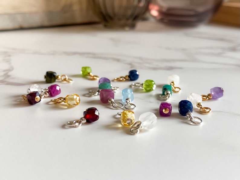 May include: Assortment of small, colorful gemstone charms with gold accents. Each charm features a different gemstone, including amethyst, aquamarine, citrine, garnet, peridot, ruby, sapphire, and topaz.
