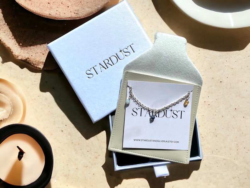 May include: A silver chain necklace with a variety of small, colorful stones hanging from it. The necklace is displayed in a white box with the word "STARDUST" printed on it.