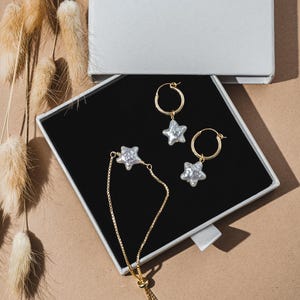 May include: A set of gold star earrings and a gold star bracelet with pearl accents. The jewellery is displayed in a white box with a black interior and the word "STARDUST" printed on the top.