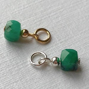 May include: Two green gemstone beads with gold and silver jump rings. The beads are faceted and have a square shape.
