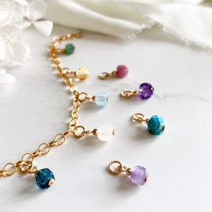 May include: A gold chain necklace with multiple charms featuring different colored gemstones, including turquoise, amethyst, aquamarine, and rose quartz.