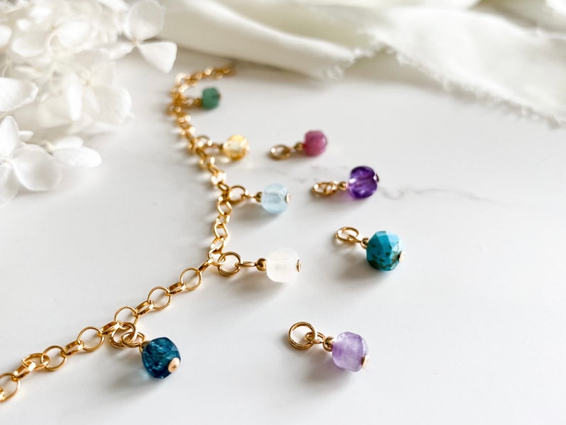 May include: A gold chain necklace with multiple charms featuring different colored gemstones, including turquoise, amethyst, aquamarine, and rose quartz.