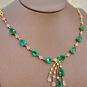 May include: A gold necklace featuring pear-shaped emeralds and small diamonds. The emeralds are a vibrant green, and the diamonds are clear. The necklace is displayed on a brown mannequin.