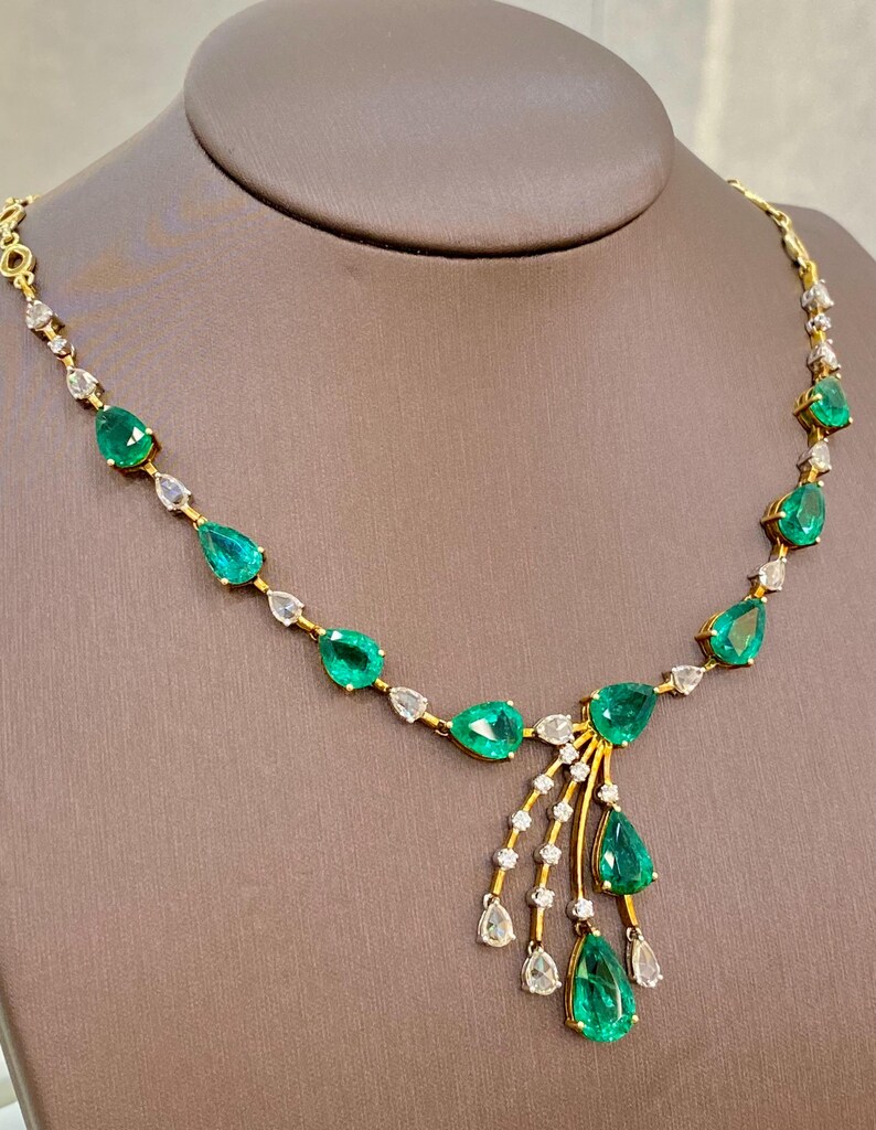 May include: A gold necklace featuring pear-shaped emeralds and small diamonds. The emeralds are a vibrant green, and the diamonds are clear. The necklace is displayed on a brown mannequin.