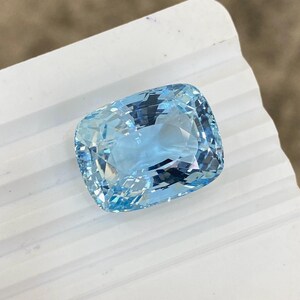 May include: A light blue aquamarine gemstone with a cushion cut. The gemstone is faceted and has a brilliant shine.
