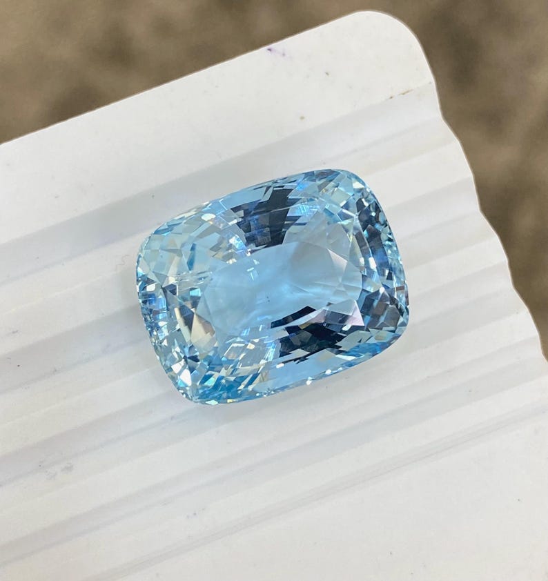 May include: A light blue aquamarine gemstone with a cushion cut. The gemstone is faceted and has a brilliant shine.