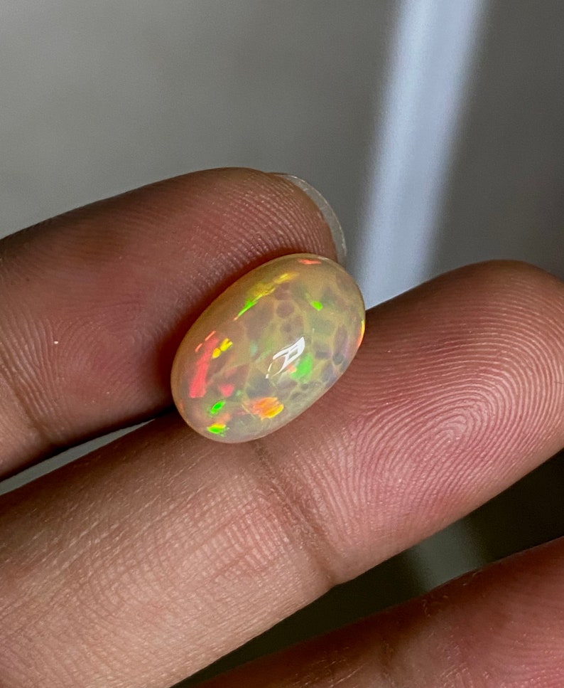 Pu&ograve; includere: Una pietra preziosa di opale ovale di colore marrone chiaro con bagliori di verde, arancione e rosso.
