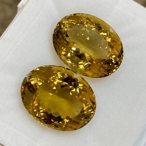 May include: Two oval-shaped, faceted, golden yellow gemstones. The gemstones are displayed on a white surface.