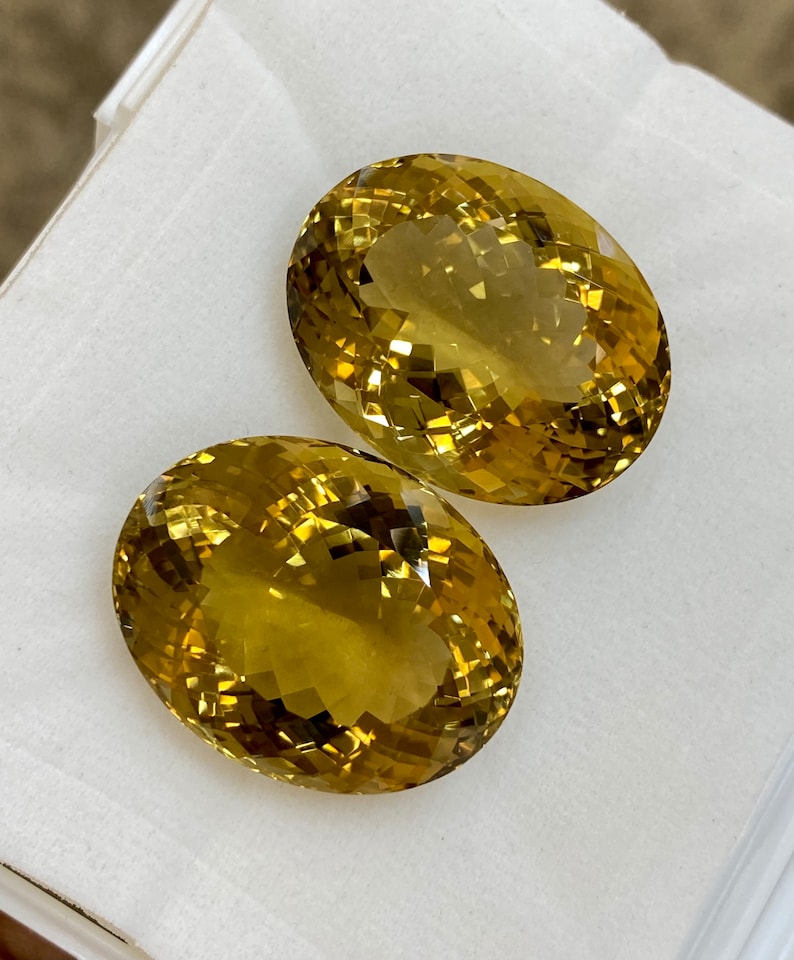 May include: Two oval-shaped, faceted, golden yellow gemstones. The gemstones are displayed on a white surface.