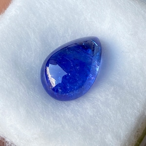 May include: A polished, teardrop-shaped, deep blue gemstone. The gem has a smooth, reflective surface and is set against a white, textured background. The stone's color is a rich, saturated blue, with hints of lighter shades visible.