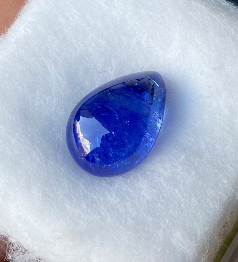 May include: A polished, teardrop-shaped, deep blue gemstone. The gem has a smooth, reflective surface and is set against a white, textured background. The stone's color is a rich, saturated blue, with hints of lighter shades visible.