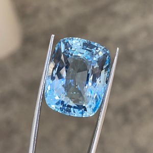 May include: A cushion-cut aquamarine gemstone with a light blue color. The gemstone is held in place by a pair of tweezers.