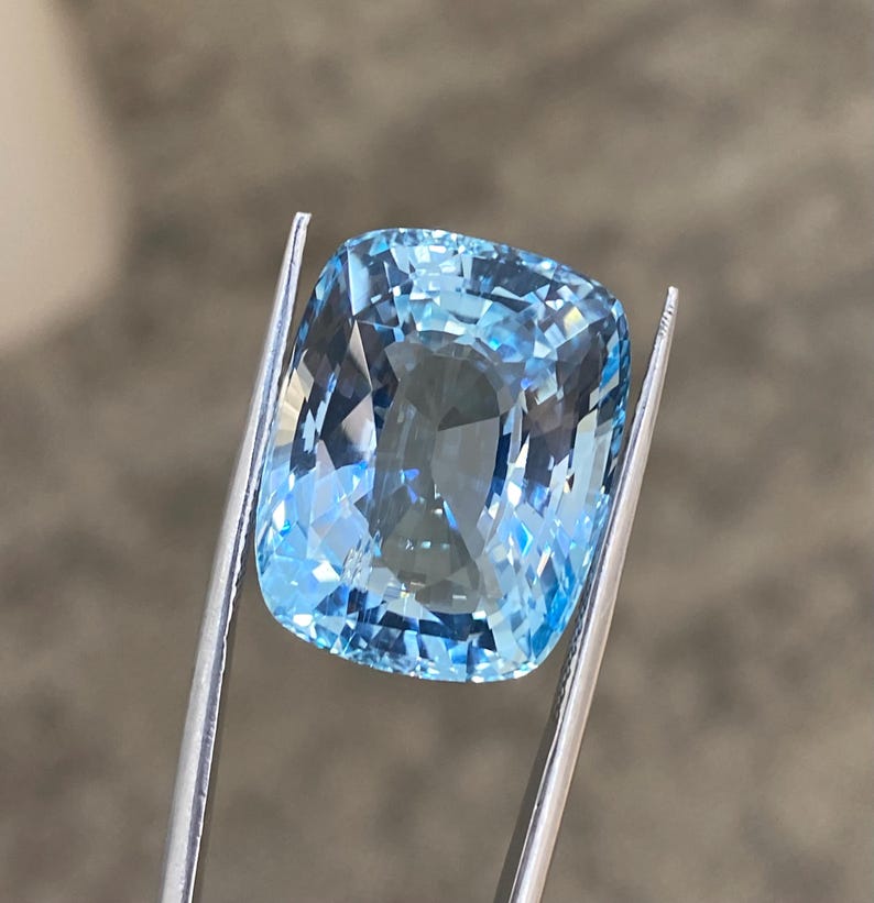 May include: A cushion-cut aquamarine gemstone with a light blue color. The gemstone is held in place by a pair of tweezers.