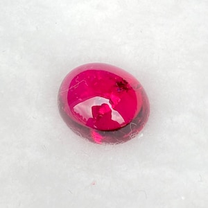 May include: A deep red oval cabochon gemstone with a smooth, polished surface.