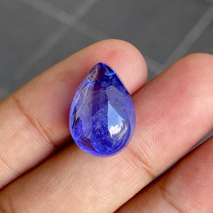 May include: A faceted, teardrop-shaped gemstone in a deep, vibrant blue hue. The gem is held between fingers, showcasing its polished surface and internal clarity. The stone's facets catch the light, creating a sparkling effect. This is a gemstone.