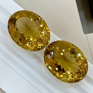May include: Two oval-shaped, faceted, golden yellow gemstones. The gemstones are on a white surface.