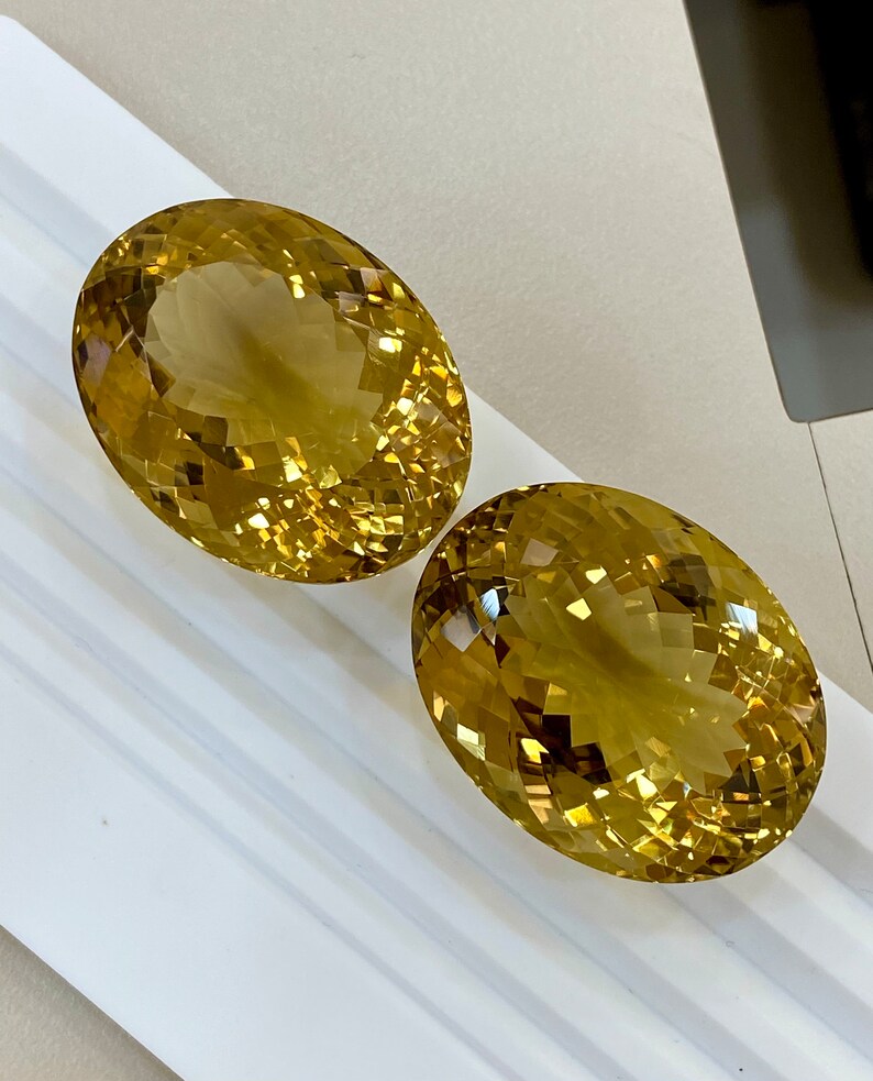 May include: Two oval-shaped, faceted, golden yellow gemstones. The gemstones are on a white surface.