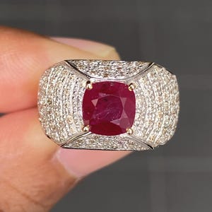 Op de afbeelding: Een ring met een vierkant geslepen robijn. De ring heeft een grote, dieprode robijn als middensteen, omringd door een halo van kleinere, sprankelende diamanten. De band is gemaakt van witgoud of platina, met een pavé-zetting van diamanten.