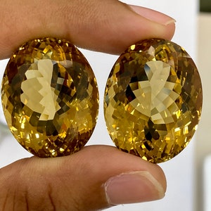 May include: Two oval-shaped, faceted gemstones with a golden yellow color. The gemstones are held in a hand.