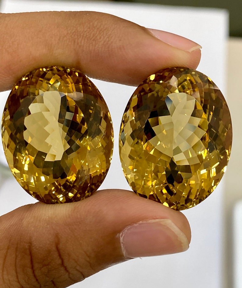 May include: Two oval-shaped, faceted gemstones with a golden yellow color. The gemstones are held in a hand.