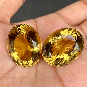 May include: Two oval-shaped, faceted citrine gemstones with a golden yellow color. The stones are set against a neutral background.