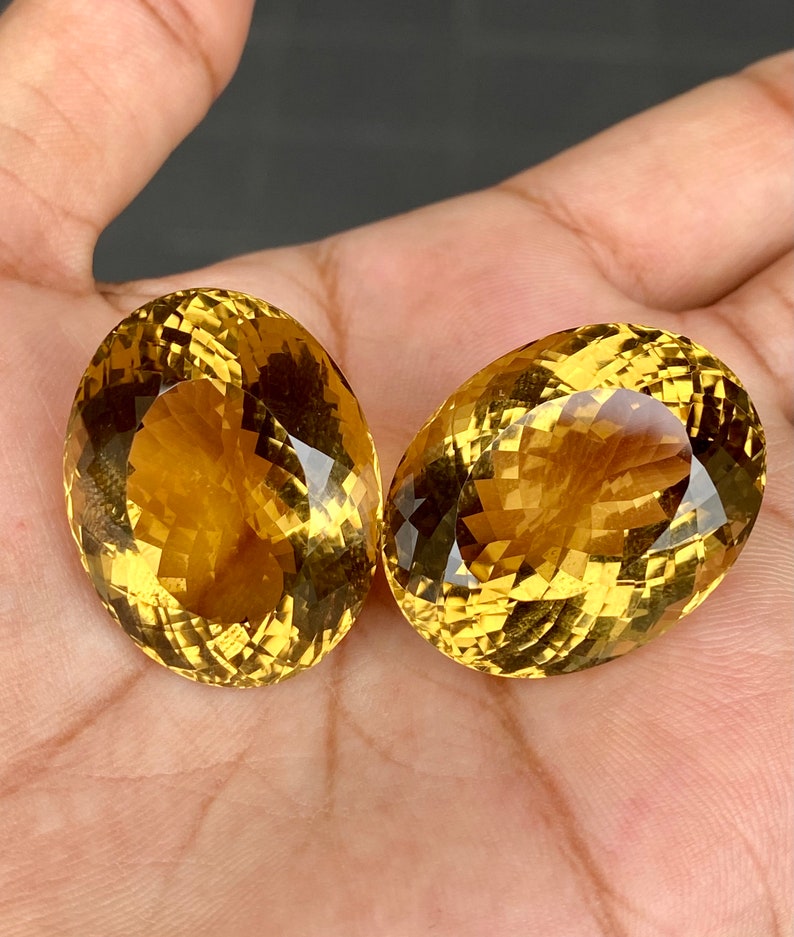 May include: Two oval-shaped, faceted citrine gemstones with a golden yellow color. The stones are set against a neutral background.
