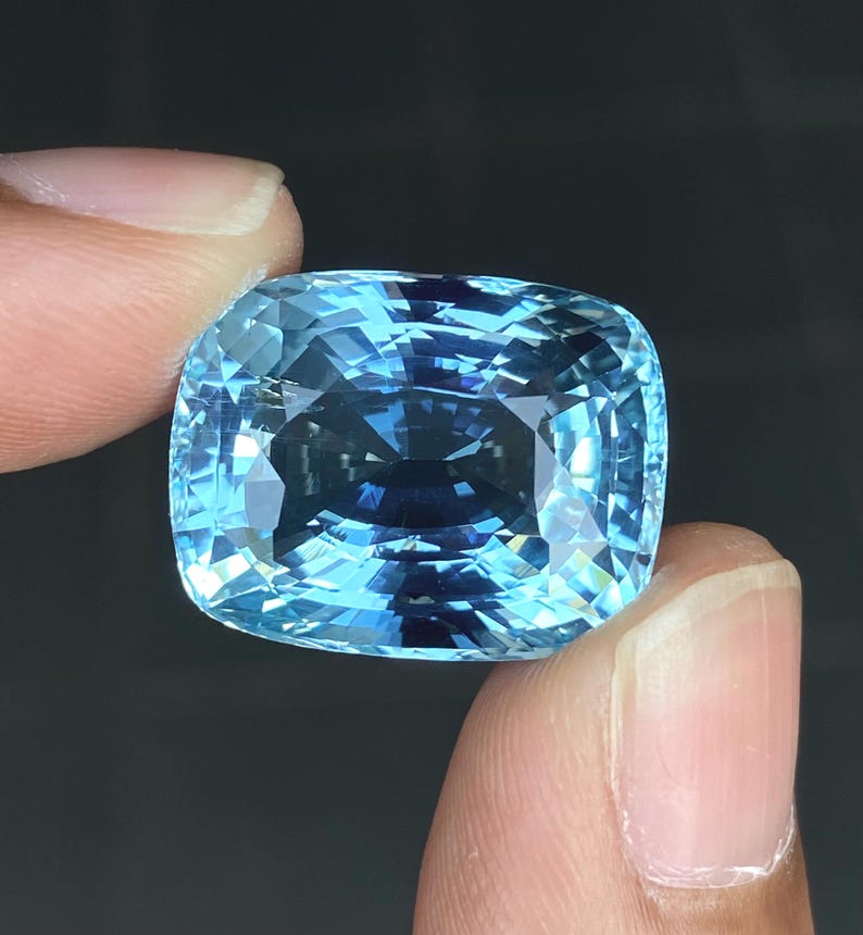 May include: A large, cushion-cut aquamarine gemstone with a light blue color. The gemstone is faceted and has a high degree of brilliance.