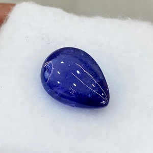 May include: A deep blue, teardrop-shaped gemstone with white inclusions and streaks. The polished surface reflects light, creating a glossy appearance. The gem is set against a white, textured background.