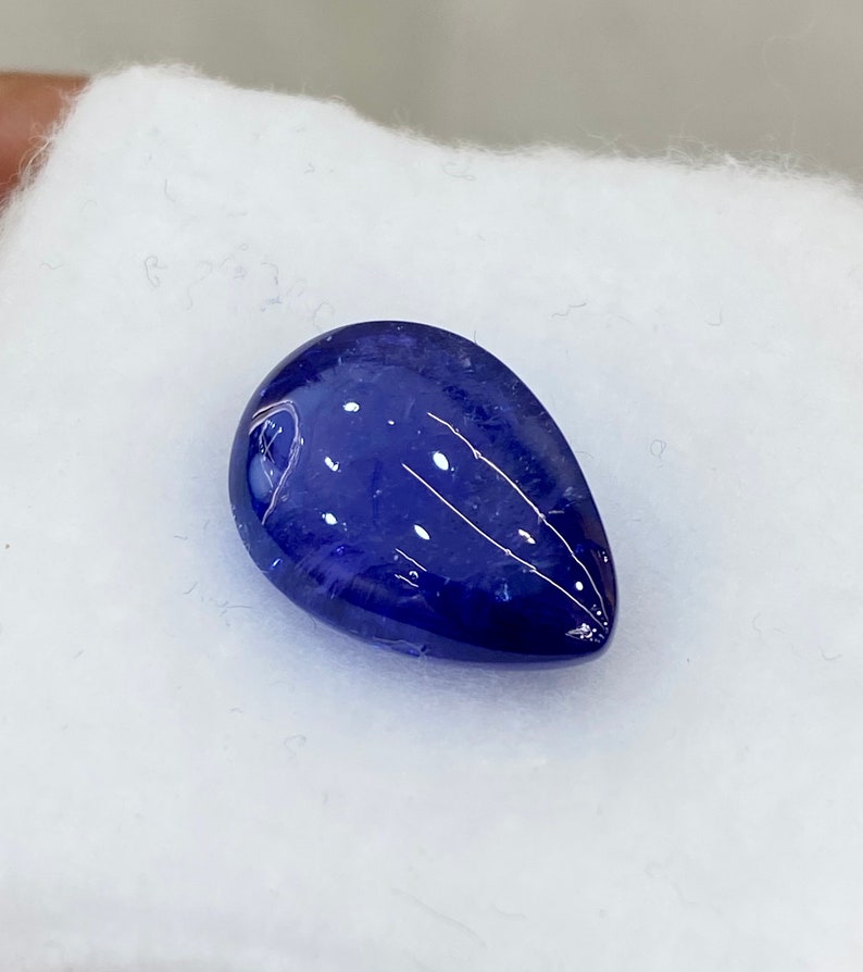 May include: A deep blue, teardrop-shaped gemstone with white inclusions and streaks. The polished surface reflects light, creating a glossy appearance. The gem is set against a white, textured background.