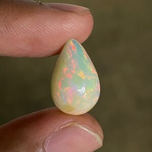 May include: A teardrop-shaped white opal gemstone with iridescent rainbow flashes. The opal is being held by a finger.