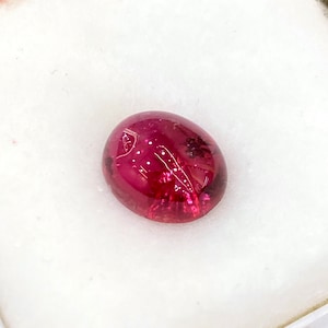 May include: A deep red oval cabochon gemstone with a smooth, polished surface.