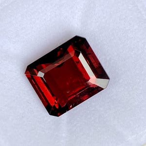 May include: A deep red gemstone with a rectangular emerald cut. The gemstone is faceted and has a high polish.