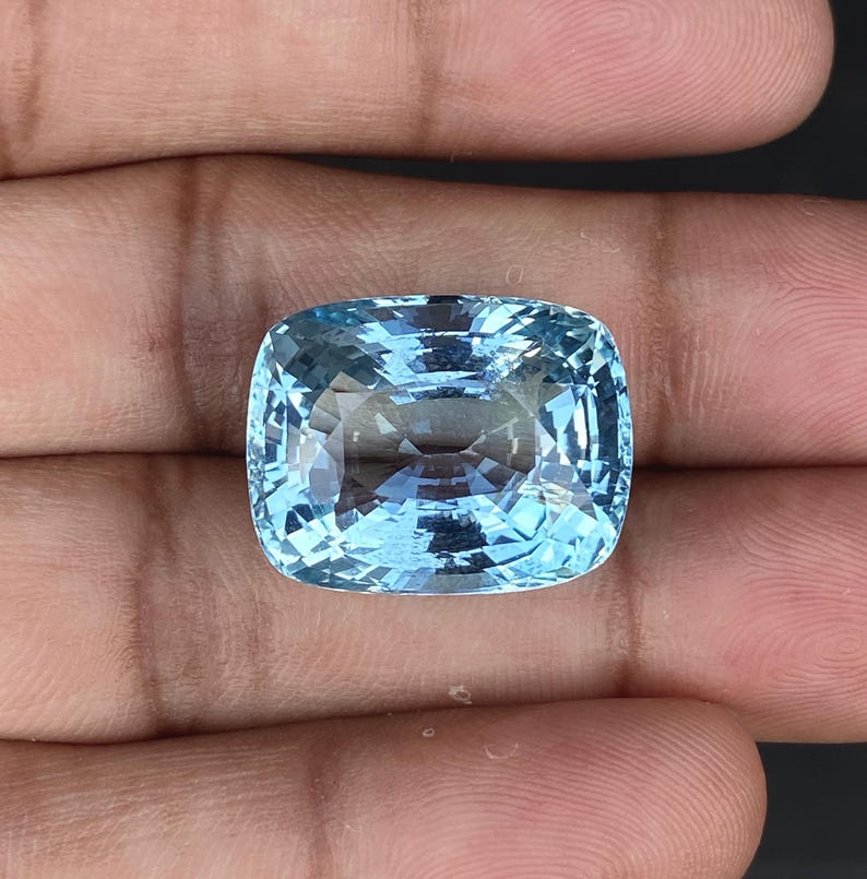 May include: A cushion-cut aquamarine gemstone with a light blue color. The gemstone is faceted and has a high degree of clarity.