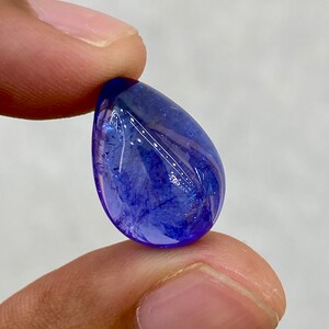 May include: A polished, teardrop-shaped gemstone with a deep blue hue. The gem has a smooth, reflective surface and internal patterns. The stone is held between two fingers against a neutral background, showcasing its color and clarity.