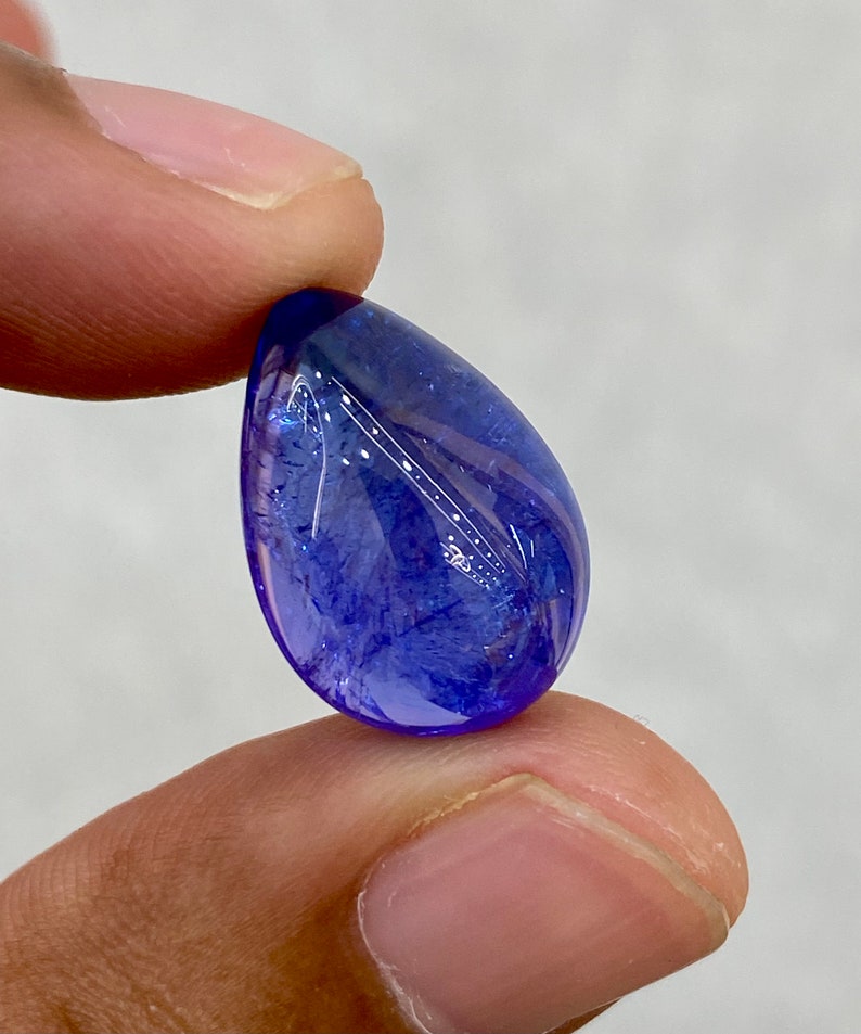 May include: A polished, teardrop-shaped gemstone with a deep blue hue. The gem has a smooth, reflective surface and internal patterns. The stone is held between two fingers against a neutral background, showcasing its color and clarity.