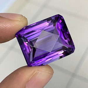 Puede incluir: Una gran gema de amatista de color púrpura intenso con un corte rectangular esmeralda. La gema está facetada y tiene un alto pulido.