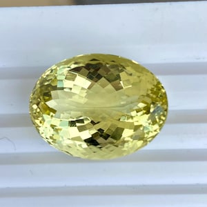 May include: A pale yellow, oval-cut gemstone with multiple facets. The surface of the gem reflects light, creating a mosaic pattern. The stone is set against a white background.