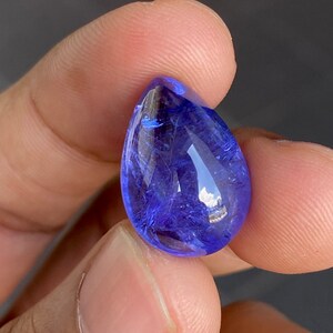 May include: A faceted, teardrop-shaped gemstone with a deep blue hue. The gem is held between fingers, showcasing its polished surface and internal clarity. The stone reflects light, highlighting its facets and color.