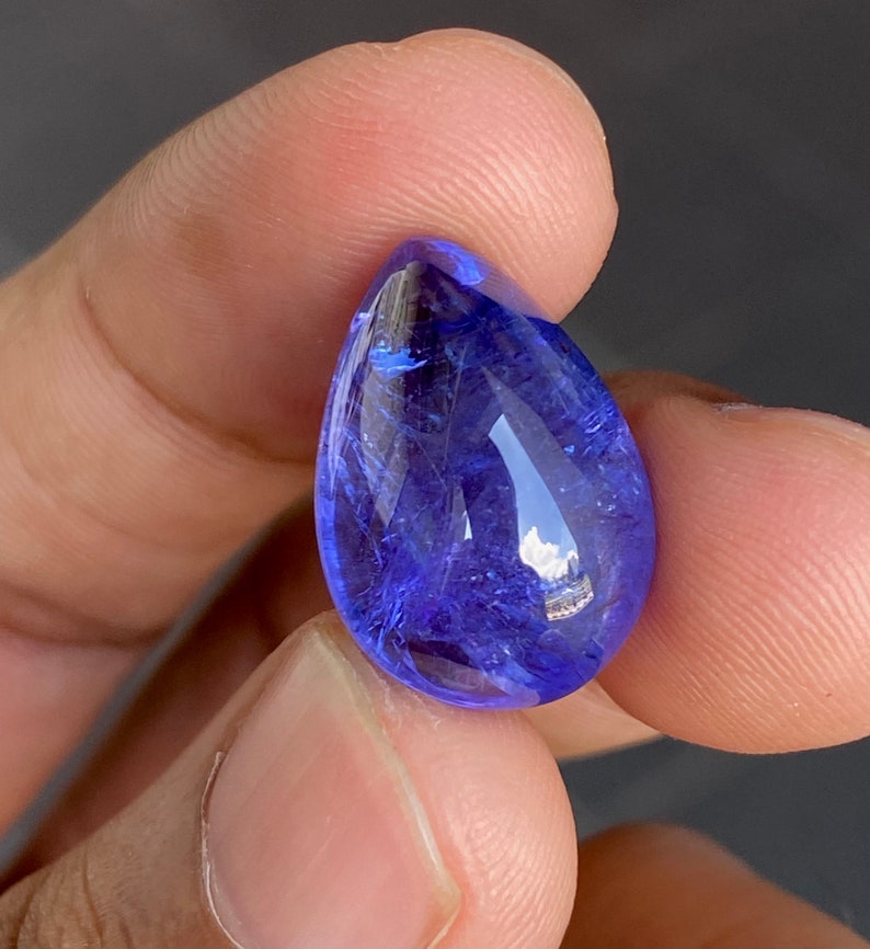 May include: A faceted, teardrop-shaped gemstone with a deep blue hue. The gem is held between fingers, showcasing its polished surface and internal clarity. The stone reflects light, highlighting its facets and color.
